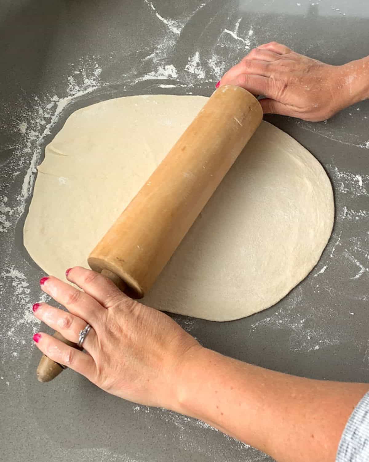 A pizza base being rolled out with a rolling pin on a lightly floured grey bench.