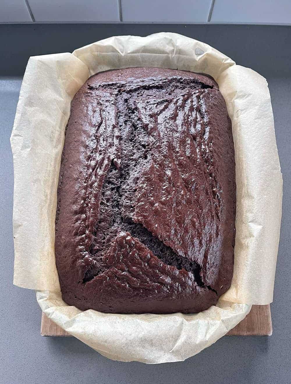 A large chocolate cake still in the tin sitting on a grey bench.