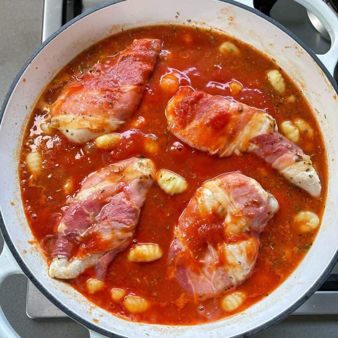 Four pieces of prosciutto-wrapped chicken simmer with gnocchi in a rich tomato sauce, creating a flavorful one pot chicken and gnocchi meal in a white pan on the stovetop.