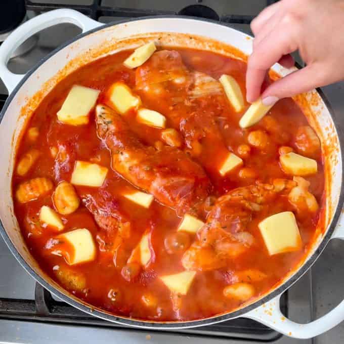 A hand adds butter pieces to a pot of one pot chicken and gnocchi simmering in tomato sauce on a stovetop.