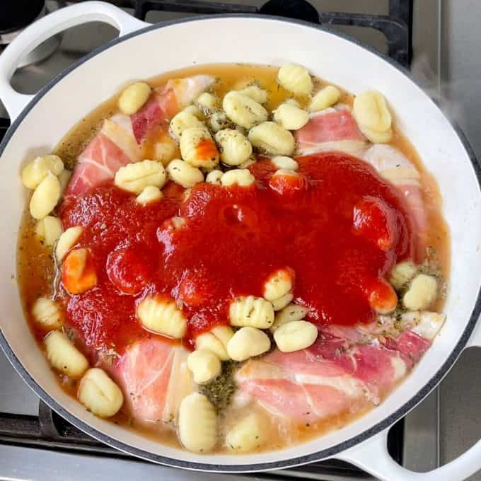 A white pan on a stove contains gnocchi, slices of ham or prosciutto, tomato sauce, herbs, and broth cooking together in a delicious one pot chicken and gnocchi-inspired meal.