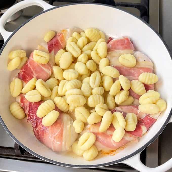 A skillet on a stove contains raw bacon-wrapped chicken pieces and uncooked gnocchi, ready to become a flavorful one pot chicken and gnocchi meal.