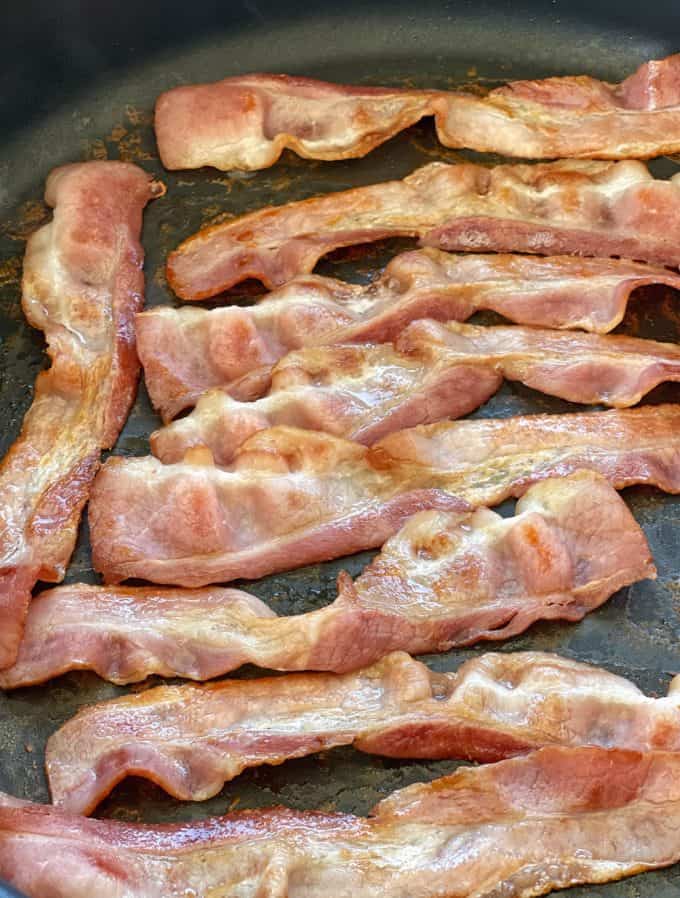 Several strips of cooked bacon are laid out in a frying pan, ready to add a savory crunch to your pesto potato salad.