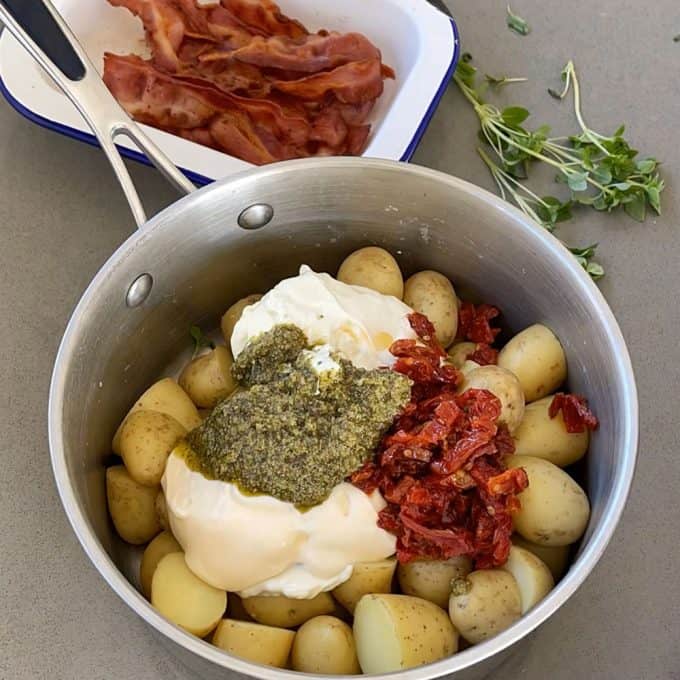 A pot of halved baby potatoes for pesto potato salad with mayonnaise, yogurt, sun-dried tomatoes, and pesto; cooked bacon and fresh herbs are nearby.