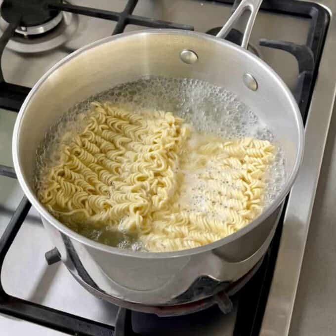 A pot of boiling water on a stove with two blocks of instant ramen noodles cooking inside, perfect for preparing a quick Chicken Satay Noodle Stir Fry.