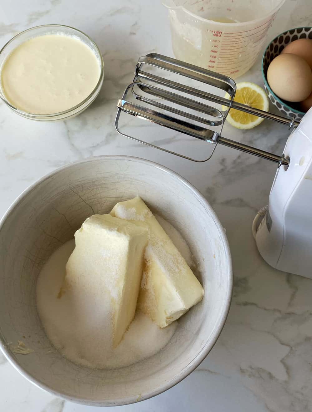 Cream cheese and sugar in a white bowl. An electric beater on its side next to the bowl with a bowl of cream, half a lemon and some eggs in the background.