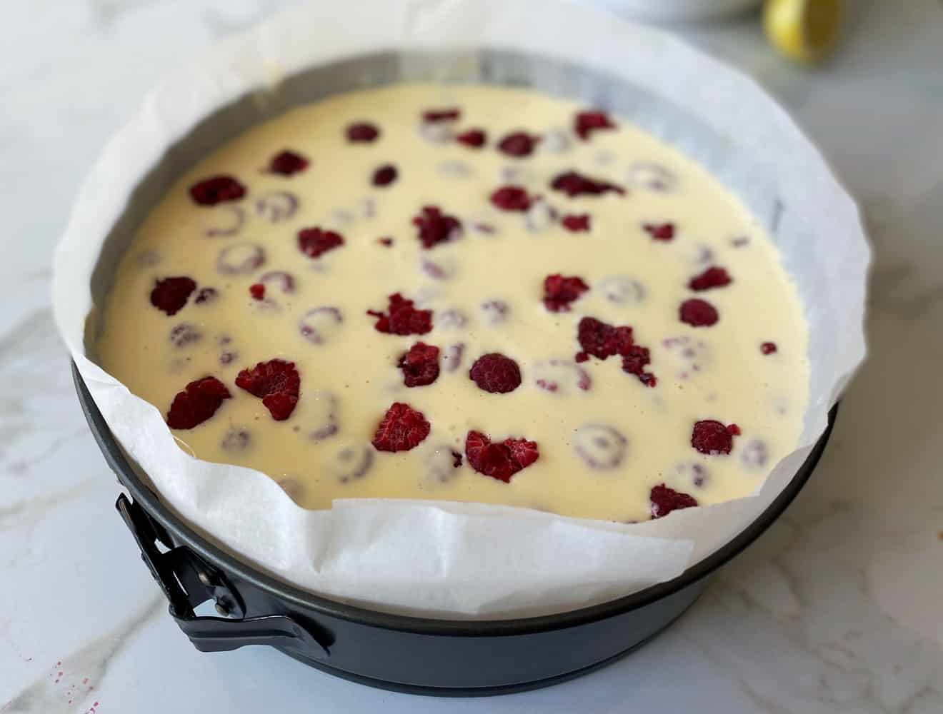 A grey, line cake tin filled with an unbaked raspberry and vanilla cheesecake on a grey and white tile.