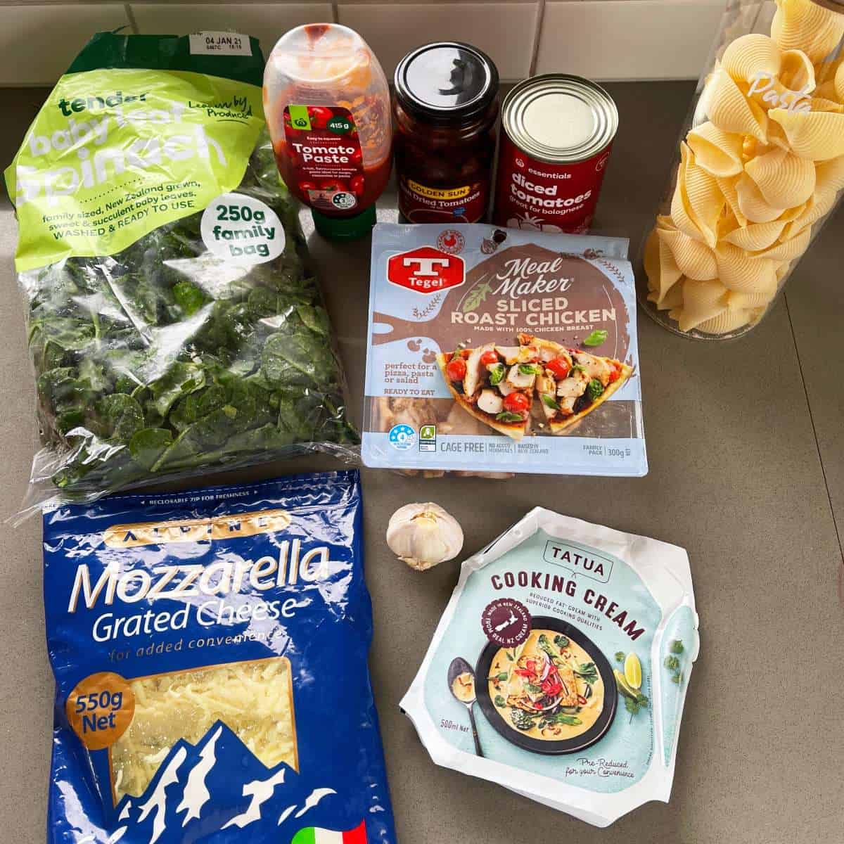A selection of ingredients on a counter for Tomato Chicken Pasta, including spinach, pasta shells, tomato paste, canned tomatoes, mozzarella cheese, cooking cream, garlic, sliced roast chicken, and chili paste.