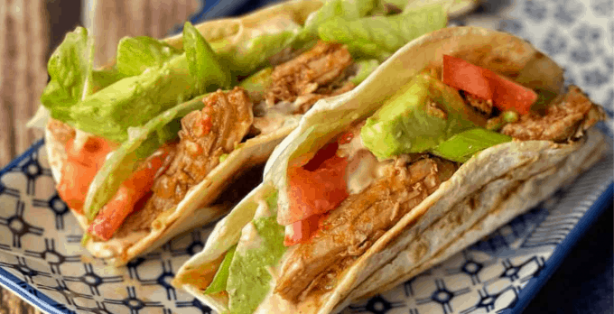 Slow Cooker Mexican Pulled Pork Two soft tacos filled with Slow Cooker Mexican Pulled Pork, sliced avocado, tomato, lettuce, and a creamy sauce are arranged on a patterned plate.