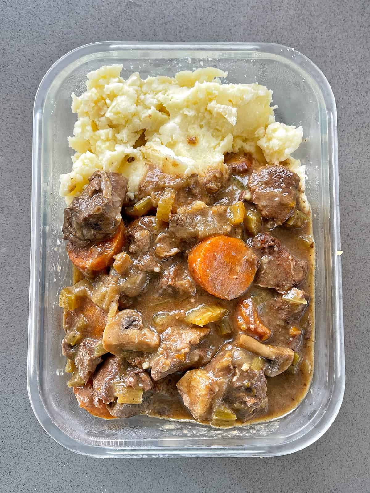 Leftover Slow Cooker Beef Stew in a glass container on a grey bench.