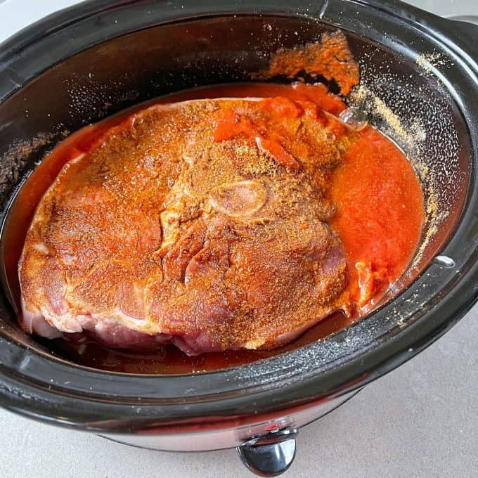 A seasoned raw pork roast sits in a slow cooker with tomato sauce and spices, ready to become flavorful Slow Cooker Mexican Pulled Pork.