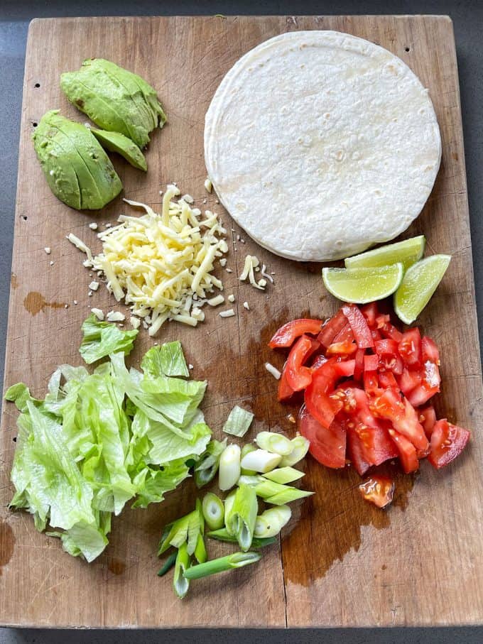 Slow Cooker Mexican Pulled Pork A wooden cutting board with two corn tortillas, Slow Cooker Mexican Pulled Pork, sliced avocado, shredded cheese, lime wedges, chopped tomato, lettuce, and sliced green onions.