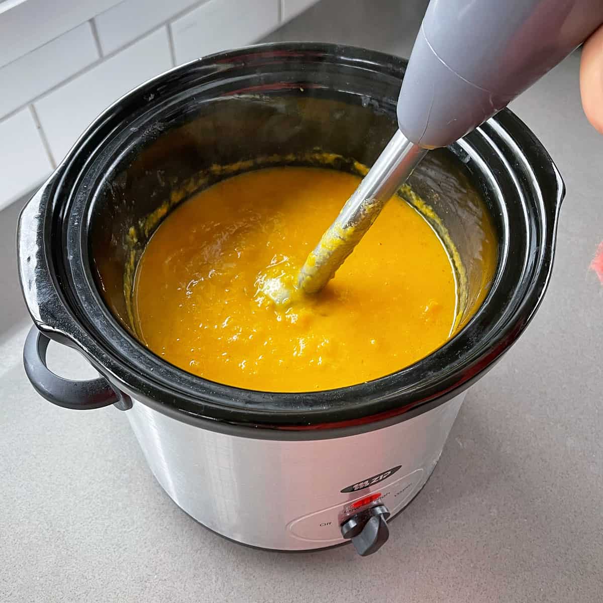 A stick blender blending pumpkin soup in a slow cooker.