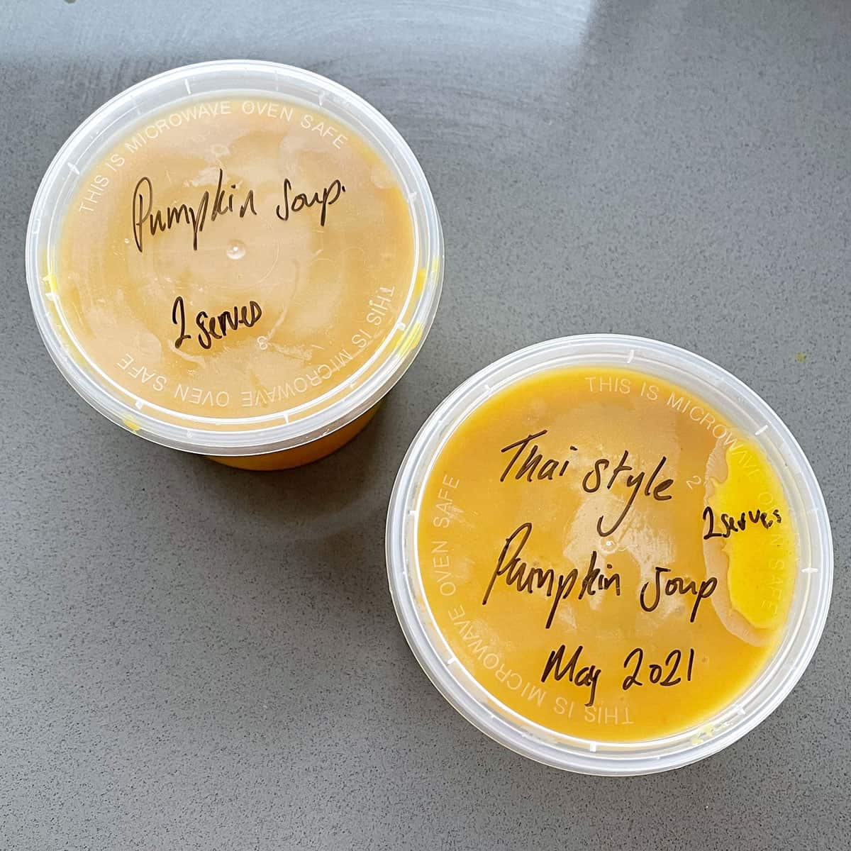 Two plastic containers of pumpkin soup sitting on a grey bench.