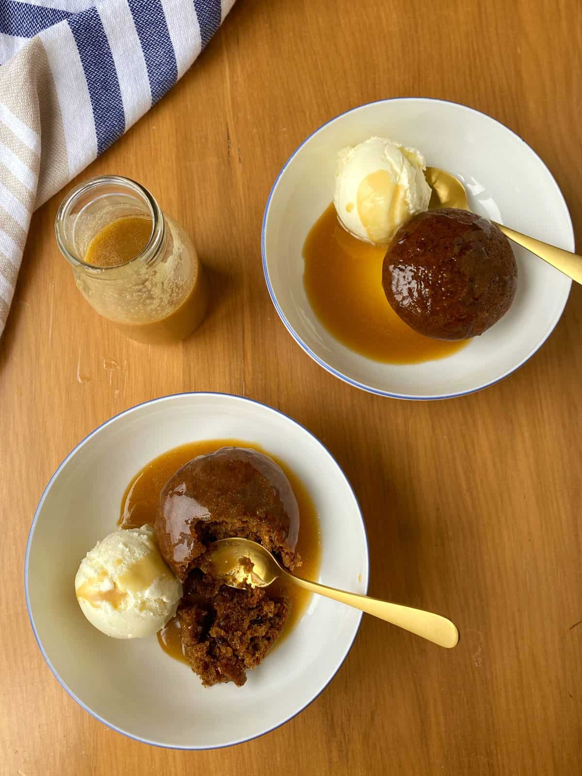 Two white bowls of sticky date pudding with butterscotch sauce and ice cream sitting on a bench.