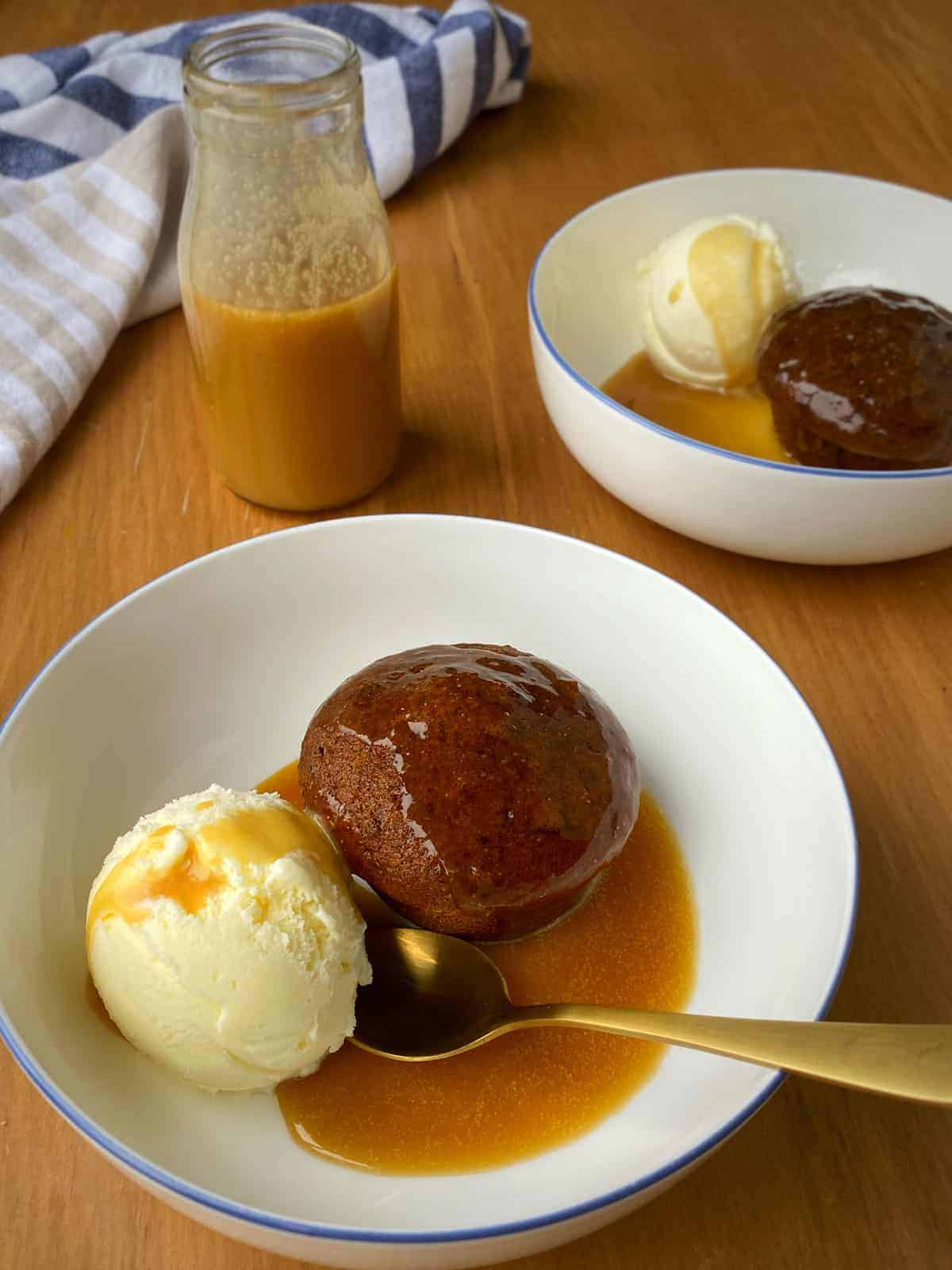 Two white bowls of sticky date pudding sitting with ice cream on a table.
