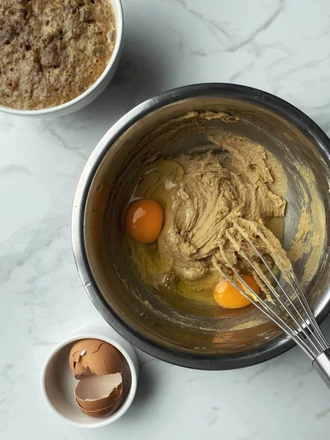 A mixing bowl with brown batter for Mini Sticky Date Puddings and two eggs, a whisk, a bowl of cracked eggshells, and a bowl of bubbling liquid on a marble surface.