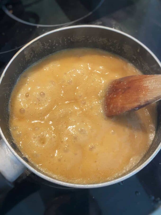 A saucepan on a stovetop with a bubbling golden brown mixture being stirred with a wooden spoon, perfect for making the rich caramel sauce for Mini Sticky Date Puddings.