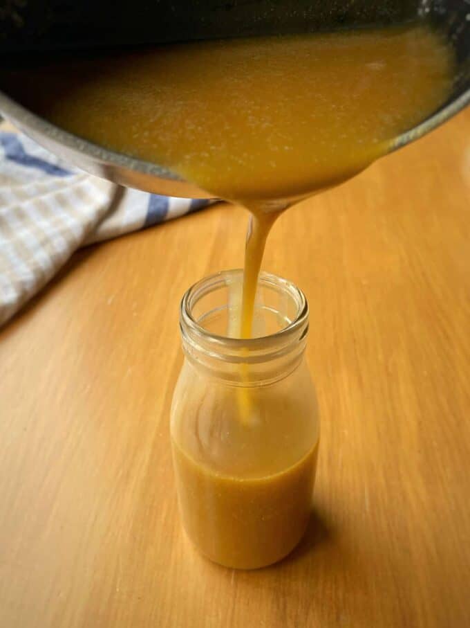 Thick golden-brown liquid, perfect for Mini Sticky Date Puddings, is being poured from a saucepan into a clear glass bottle on a wooden surface.
