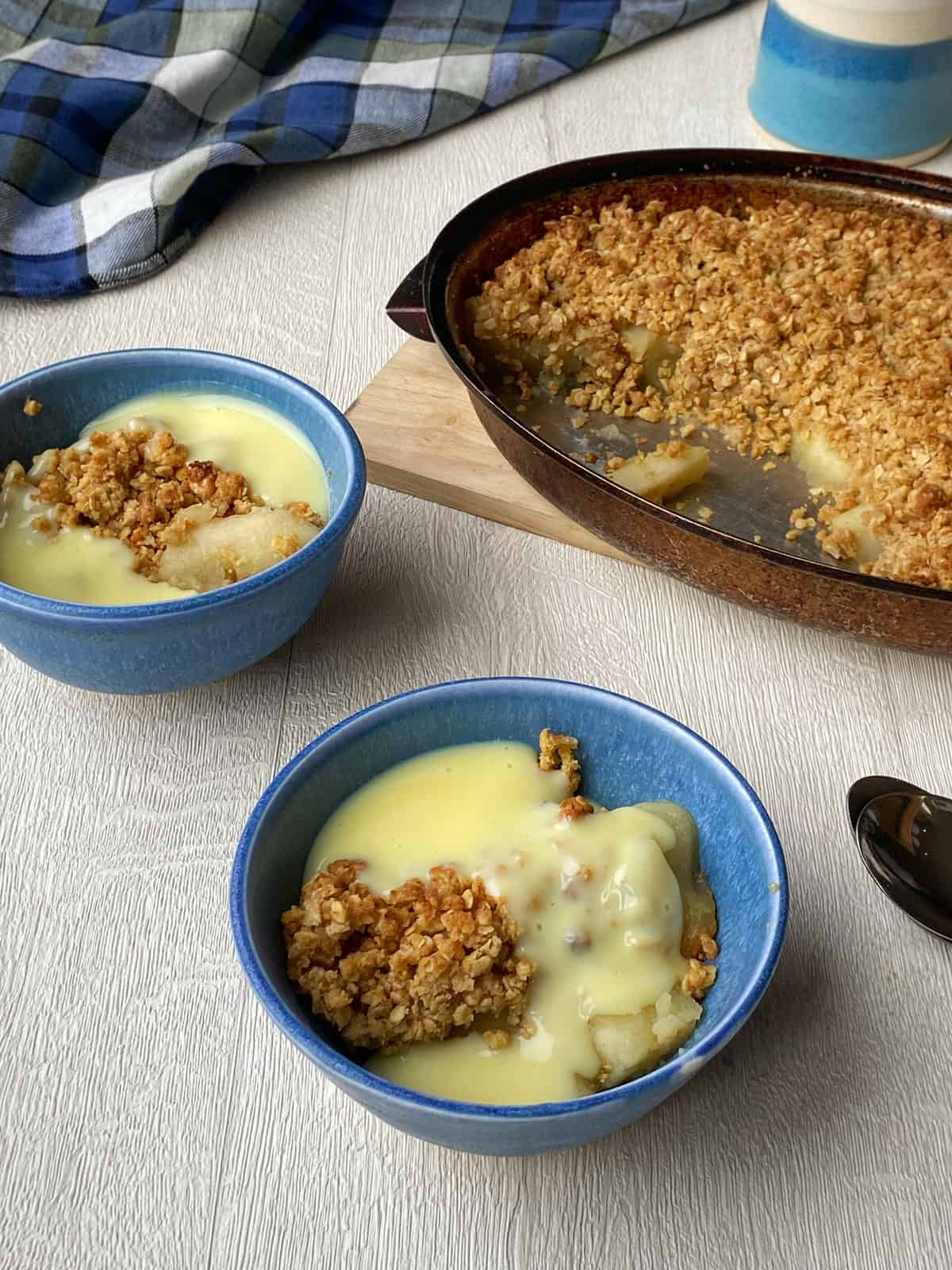 a large dish of apple crumble and two smaller bowls of apple crumble with custard sitting on a white bench.