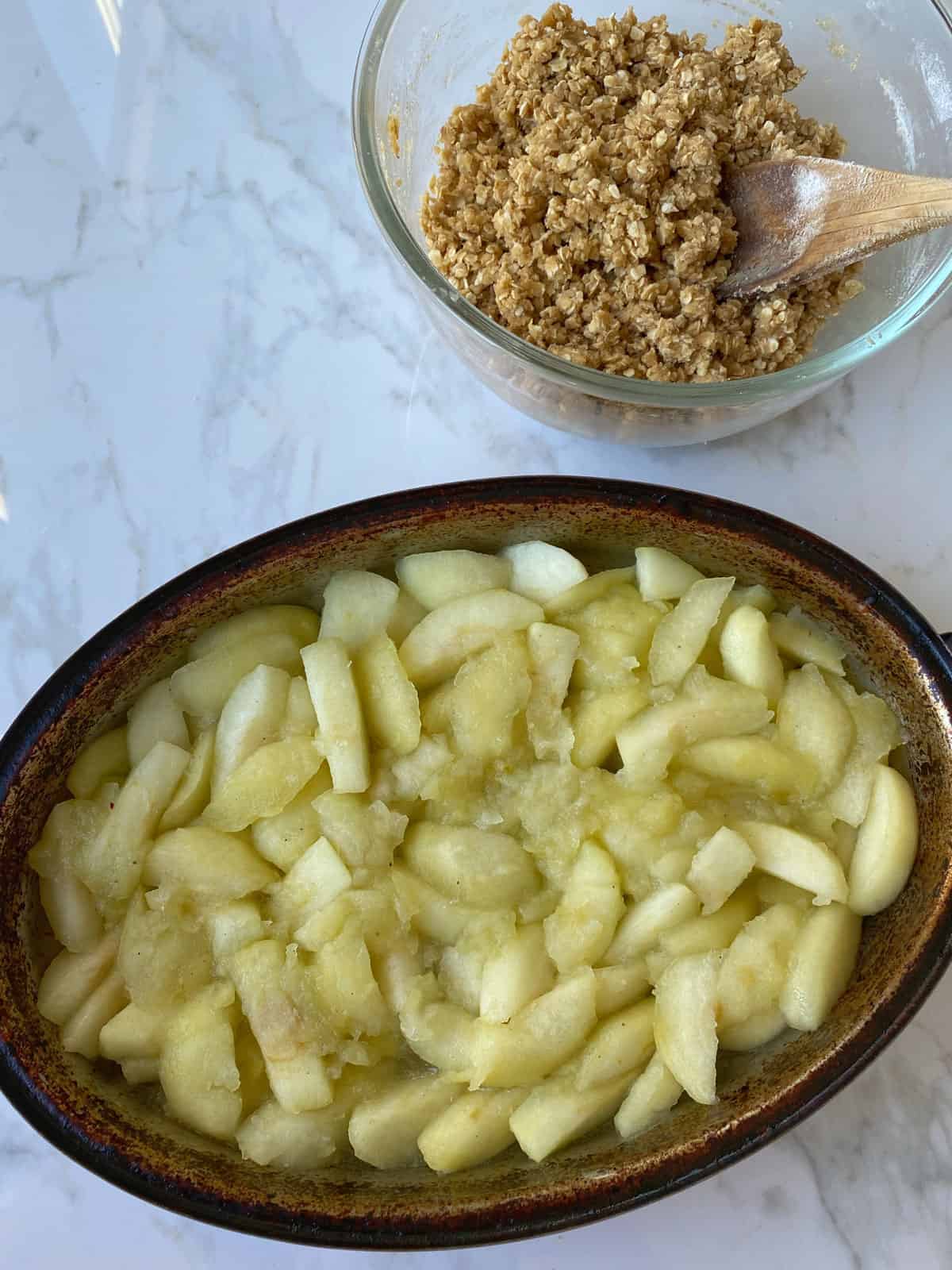 Cooked apple pieces in an oval dish and crumble topping in a glass bowl on a white bench.