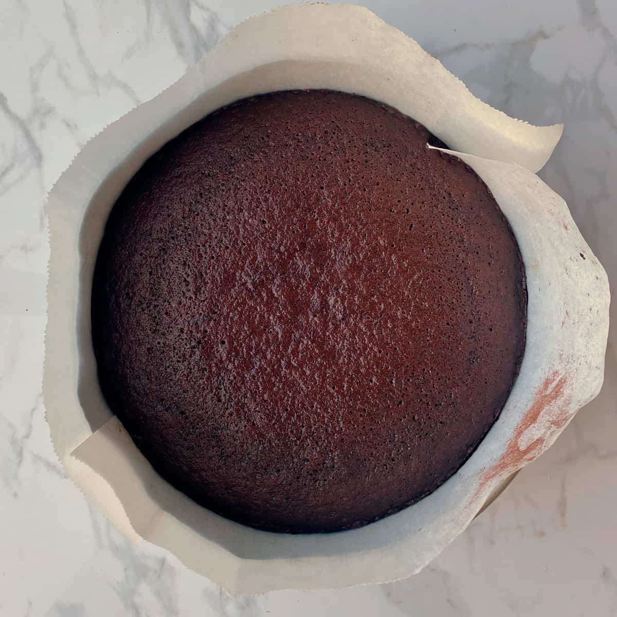 Baked chocolate cake in a lined cake tin on a bench.