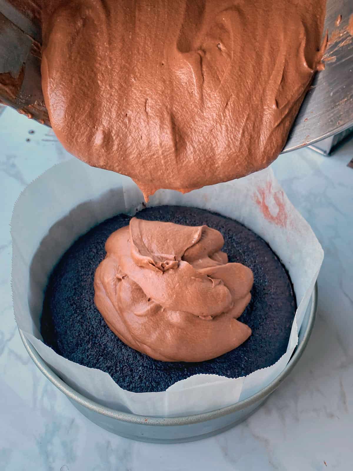 Chocolate mousse being poured onto a chocolate cake in a tin.