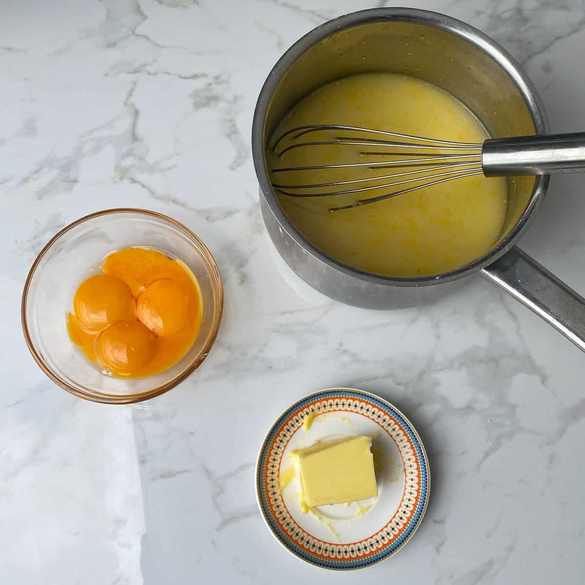 Egg yolks in a bowl, butter on a plate and a lemon mix in a saucepan all on a bench.