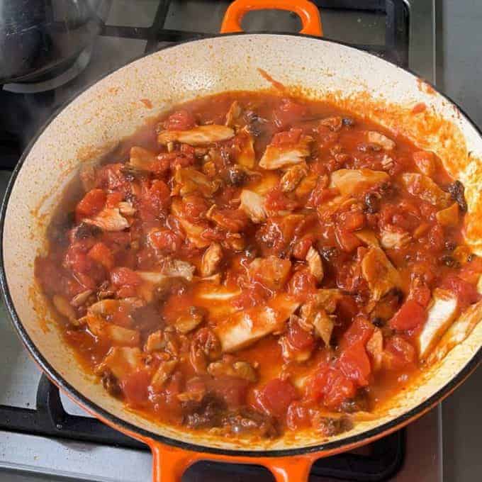 Tomato-based sauce with pieces of cooked fish simmers in an orange cast iron pan on a stovetop, perfect alongside Tomato Chicken Pasta.