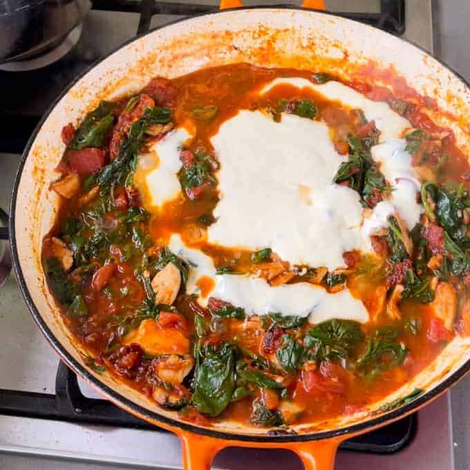 A skillet on a stovetop containing a hearty tomato chicken pasta stew with spinach, mushrooms, and a dollop of white sauce or yogurt on top.