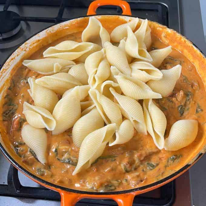 Large pasta shells are nestled on a creamy tomato-based sauce with spinach, mushrooms, and tender chicken in an orange pan on the stovetop for a comforting Tomato Chicken Pasta dish.
