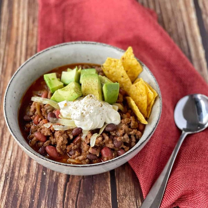 Chilli con carne served with avocado, sour cream and nacho chips in a white bowl.
