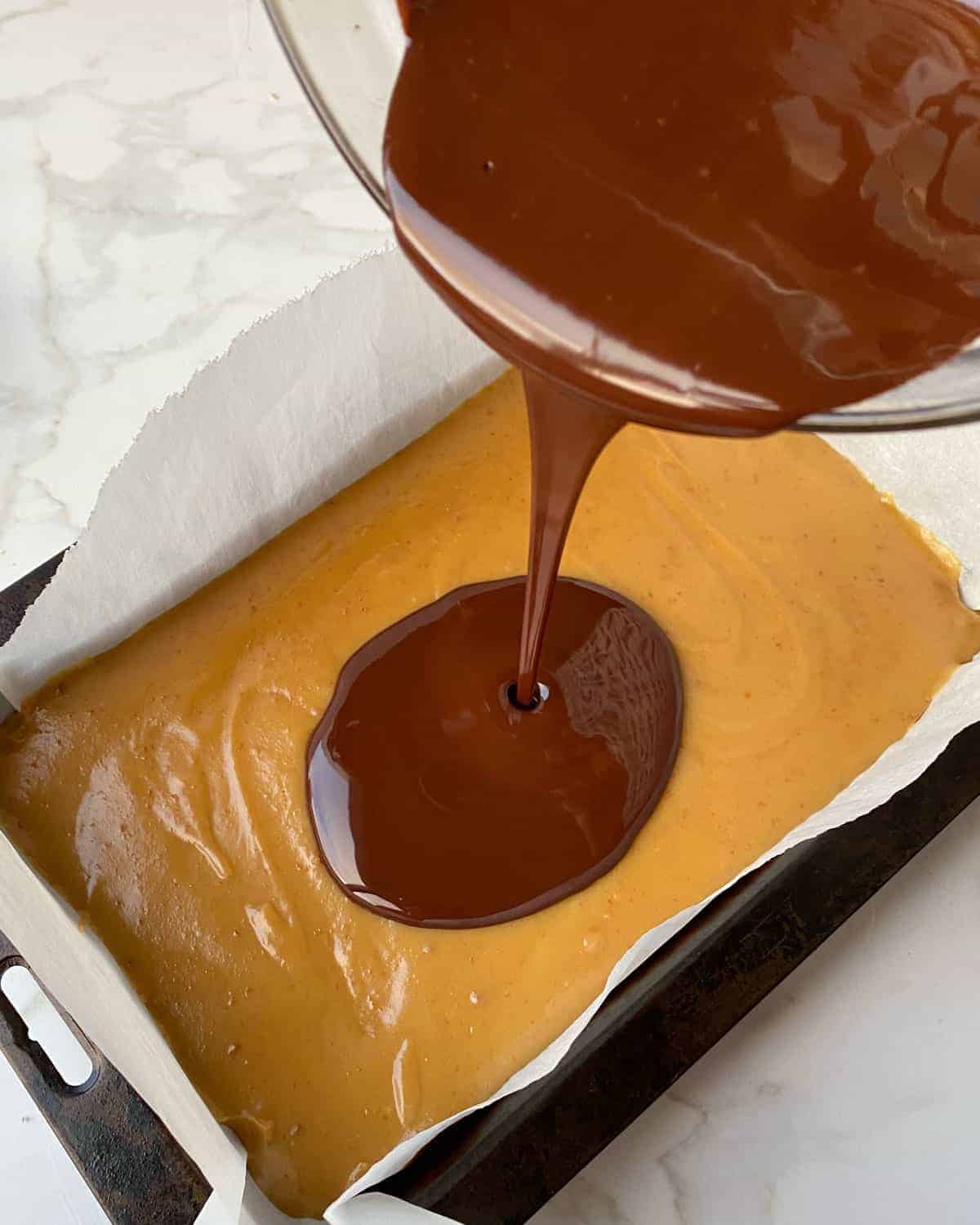 Chocolate being poured onto caramel in a lined baking tray.