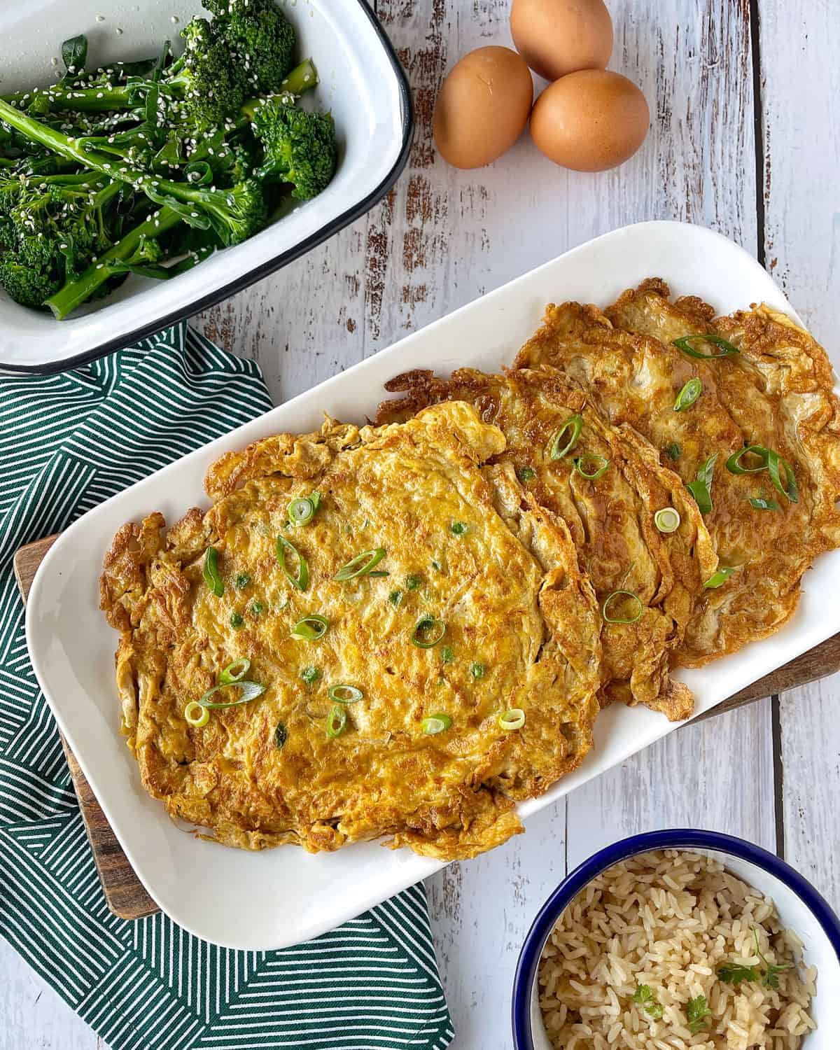 Egg Foo Young on white plate surrounded by fresh eggs, cooked broccoli and rice.