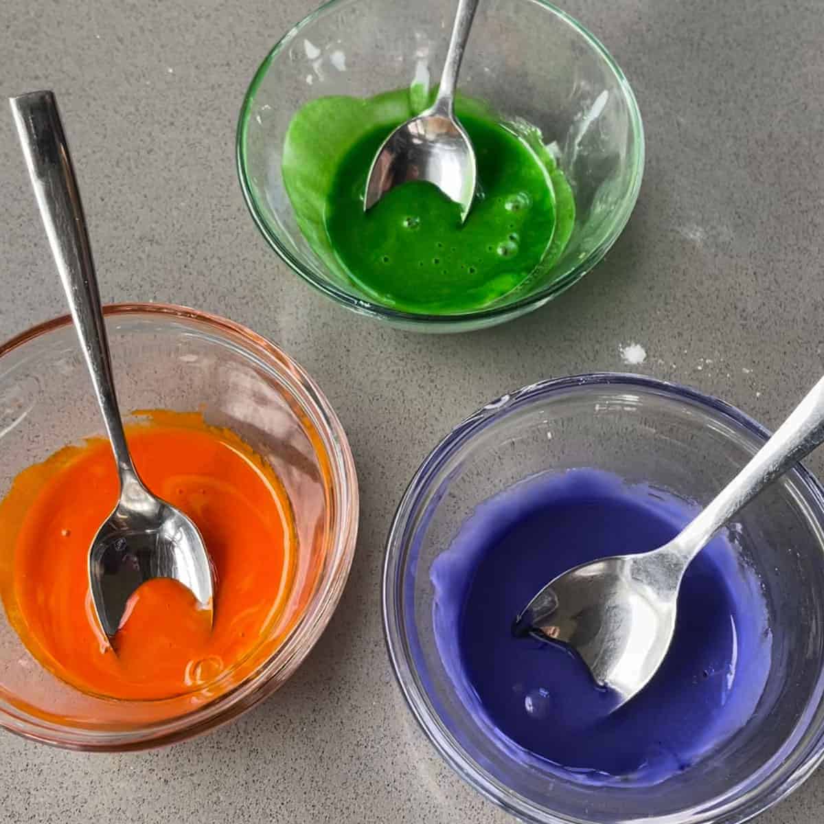 Green, orange and blue icing in glass bowls on a grey bench.