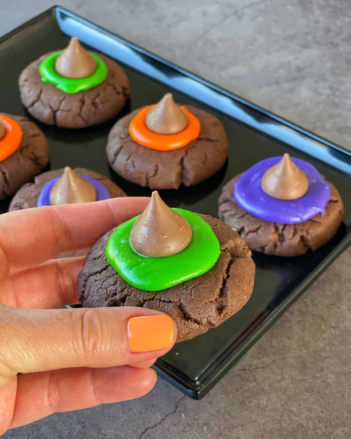 A hand holding a witches hat cookie with a tray of cookies in the background.