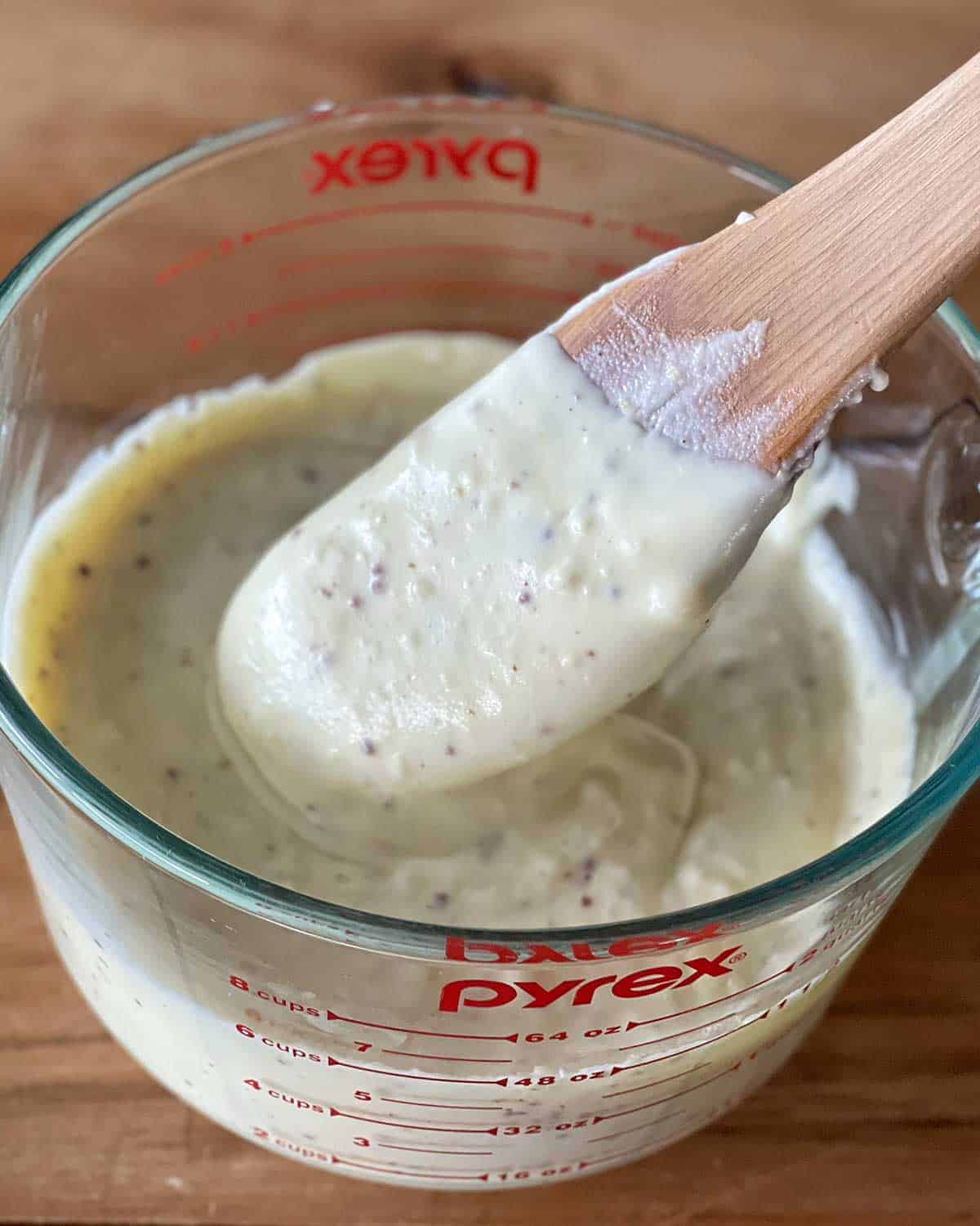 A close up of white sauce in a glass jug being stirred by a wooden spoon.