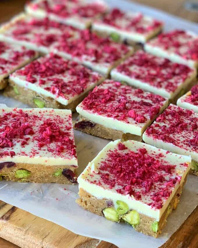 Squares of cranberry and pistachio slice with a white topping, sprinkled with crushed freeze-dried raspberries and visible pieces of pistachio, rest on a sheet of parchment paper.