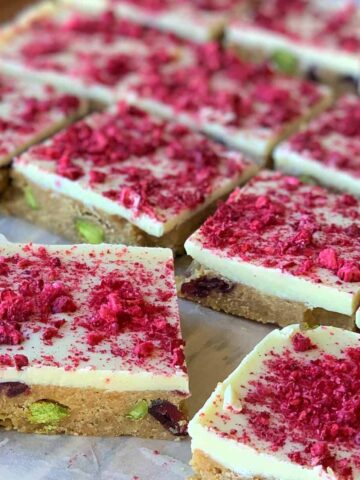 Squares of cranberry and pistachio slice with a white topping sprinkled with red crumbs, set on a brown base filled with visible nuts and fruit pieces, arranged in rows on parchment paper.