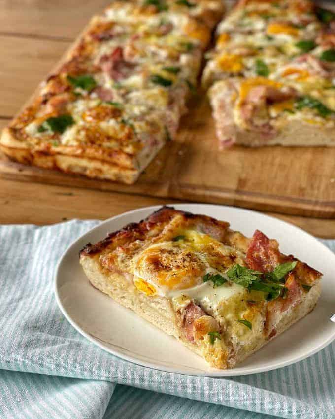 Breakfast Pizza Slab A square slice of Breakfast Pizza Slab with egg, ham, cheese, and herbs on a plate, with more slices on a wooden board in the background.