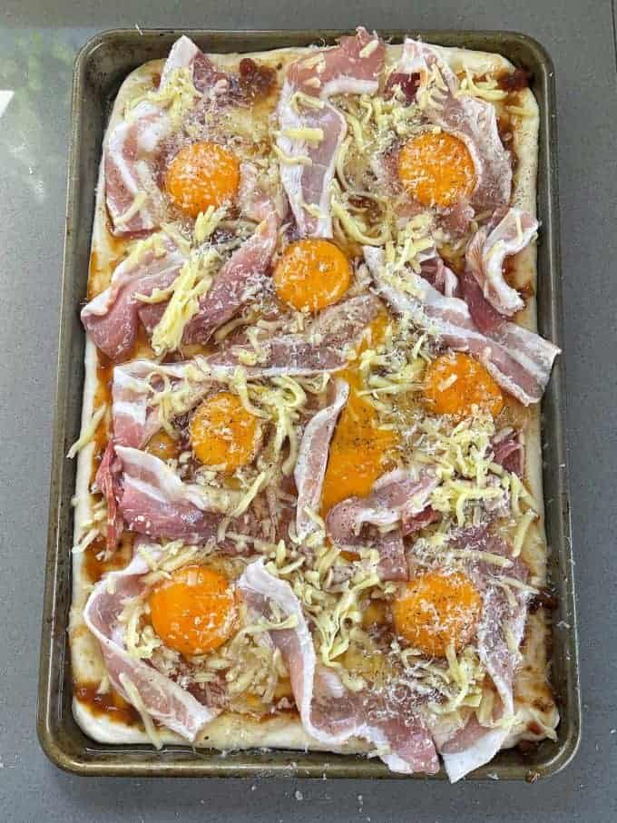 A rectangular baking tray with dough topped with raw bacon, grated cheese, raw egg yolks, and seasoning—this Breakfast Pizza Slab is ready to be baked.