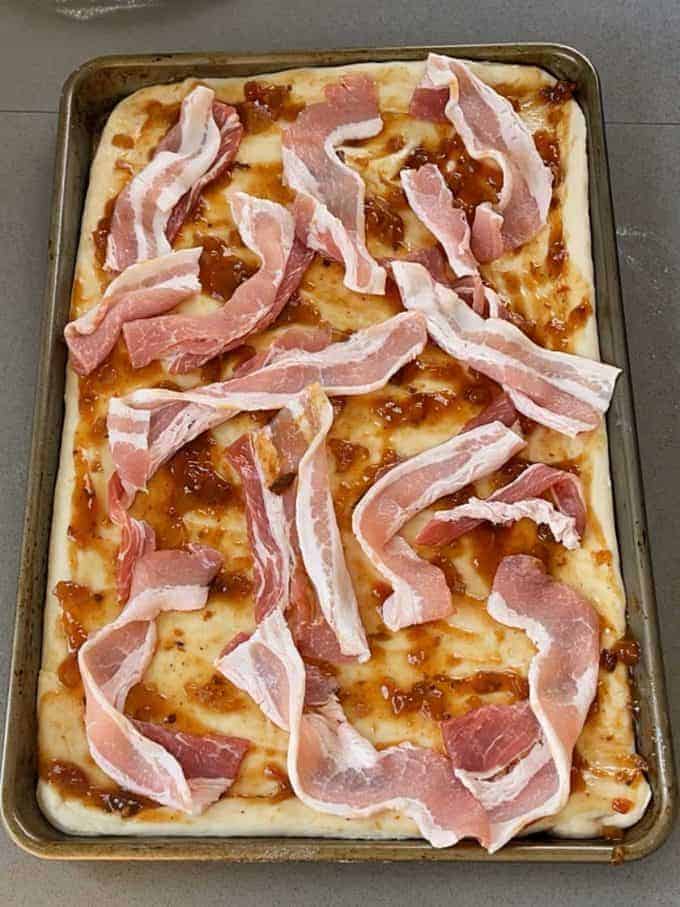 Breakfast Pizza Slab featuring raw pizza dough on a baking sheet, topped with sauce, cheese, and uncooked strips of bacon—ready to be baked to perfection.