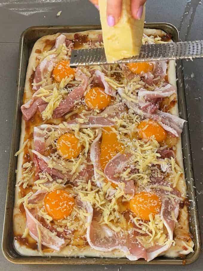 Breakfast Pizza Slab A hand grates cheese over a Breakfast Pizza Slab featuring raw eggs, shredded cheese, and slices of prosciutto on a rectangular baking sheet.
