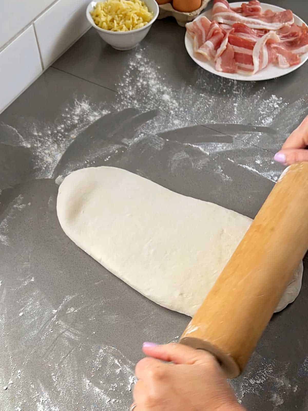 Pizza dough being rolled out on a floured, grey bench.