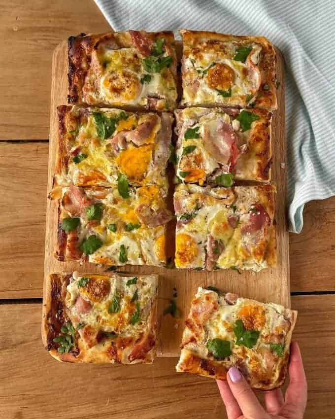 Breakfast Pizza Slab A rectangular pizza on a wooden board, cut into nine square slices with visible eggs, bacon, cheese, Belgian Biscuits crumbs, and herbs; a hand lifts one slice.