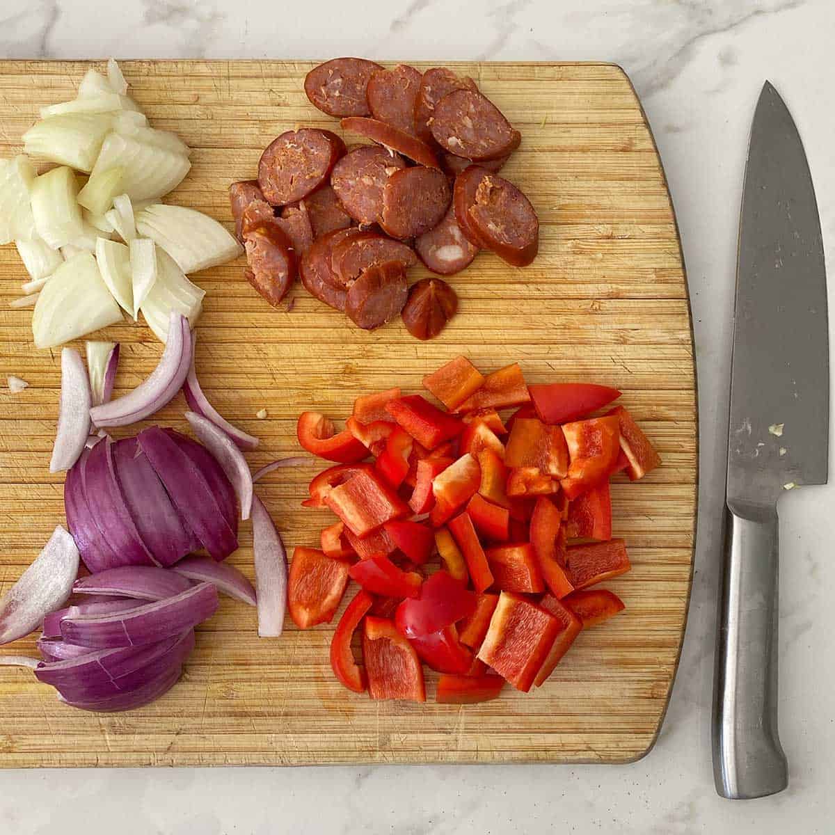 Onion, chorizo and capsicum on a wooden chopping board next to a large knife.