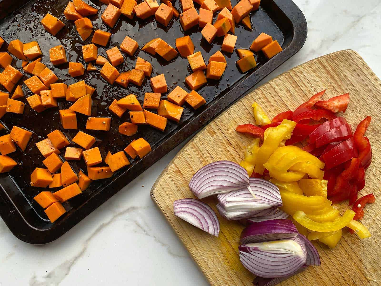 Pumpkin pieces on a baking tray next to sliced capsicum and red onion on a wooden chopping board.