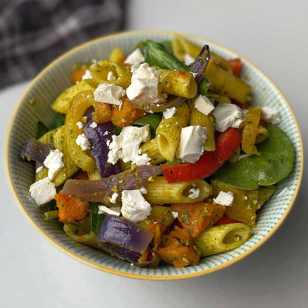 A close up of Roasted vegetable and pesto pasta salad in a white bowl on a bench.
