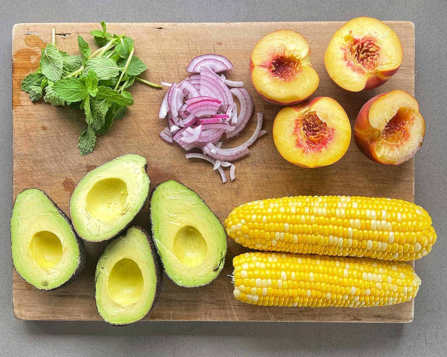 Avocado, mint, red onion, peach and corn on a wooden chopping board.