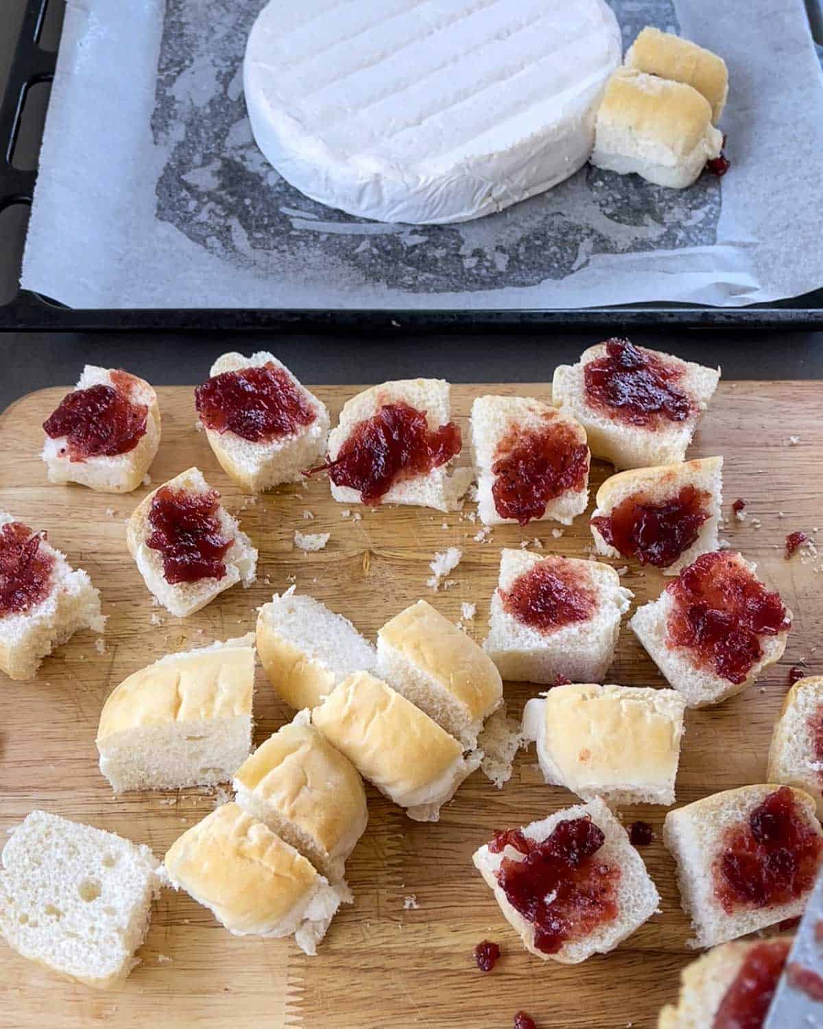 A large cog of brie on a lined baking tray next to bread rolls spread with cranberry sauce on a wooden chopping board.